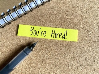 You're Hired written on yellow sticky notes next to a black pen and notebook. 