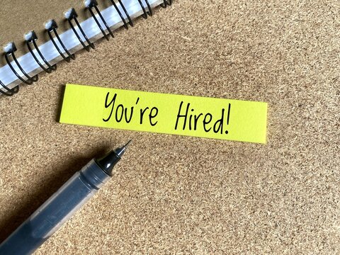 You're Hired written on yellow sticky notes next to a black pen and notebook.  - Powered by Adobe