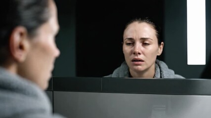 A woman sits in a shadowy room, wearing a robe. She gazes into the mirror with intense focus, showing a range of emotions from contemplation to concern as she confronts her inner thoughts.