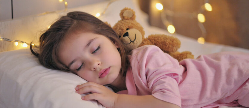 Dreamland slumber: A peaceful portrait of a young girl embraces sleep, snuggled beside a cuddly teddy bear, serenaded by the soft glow of fairy lights.