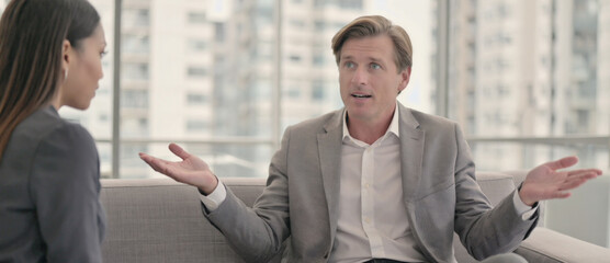 Business Discussion: A male executive in a gray suit gestures with both hands in a business setting, his words shaping a focused conversation.