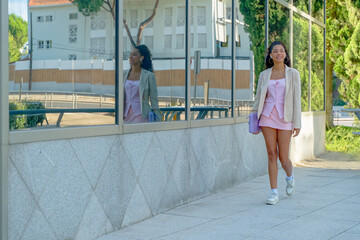 Young woman confidently walking on a city sidewalk while reflecting in a building's window