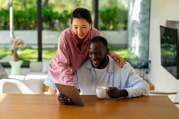 Happy young diverse, multiethnic couple planning budget, reading good news on digital tablet at home