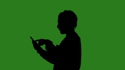 A black isolated silhouette of a man standing in profile with a smartphone in his hands, against a green chroma key background. Minimalistic contrast and modern visual composition, mockup.