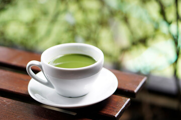 White cup of green tea on wooden table with green nature background.