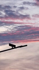 modern street light arm and its housing silhouetted in black against a vibrant sunset sky. The sky is filled with striated, high-altitude clouds illuminated in vivid pink, purple, and pale blue hues, 