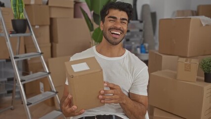 Man holds cardboard box smiling among stacked moving boxes and ladder in building; joy homecoming.