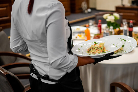 A waiter in an upscale restaurant wearing a white shirt, black apron, and gloves, serving two plates of Olivier or Russian salad at a set dining table for a formal event - Powered by Adobe