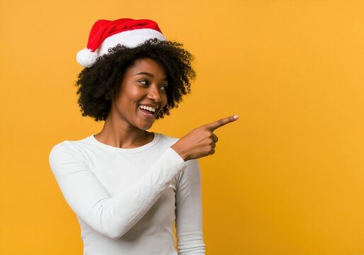 Happy young Black woman in a Santa hat smiling and pointing. Cheerful African American female model showing something for a Christmas holiday sale isolated on a yellow background with copy space