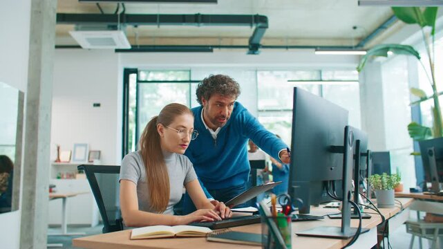 Curly-haired male manager holding clipboard standing beside focused female employee typing on keyboard. Team correcting error in financial data together in bright organized office setting.