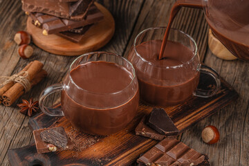 Hot chocolate being poured into glass mugs with pieces of dark and milk chocolate, cinnamon sticks, and hazelnuts on rustic wooden background, cozy winter beverage concept