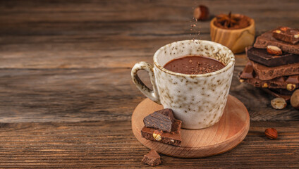 Cup of hot chocolate on wooden table with falling cocoa powder, pieces of dark and milk chocolate with hazelnuts, cozy rustic background with copy space for warm drink concept