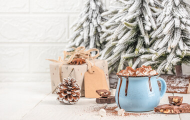 Cup of hot chocolate with marshmallows in blue mug on wooden table with Christmas decorations, pine cone, gift box and snowy fir trees, cozy winter holiday scene with copy space
