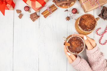 Top view of woman holding cup of hot chocolate with cinnamon on light wooden background surrounded by gifts, candy canes, and Christmas decorations, cozy winter holiday scene with copy space