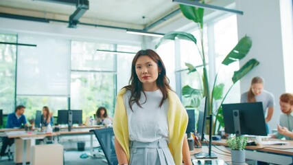 Smiling Asian woman standing confidently with arms crossed in open office. Warm friendly expression. Colleagues working at desks in background in bright collaborative workspace with green plants.