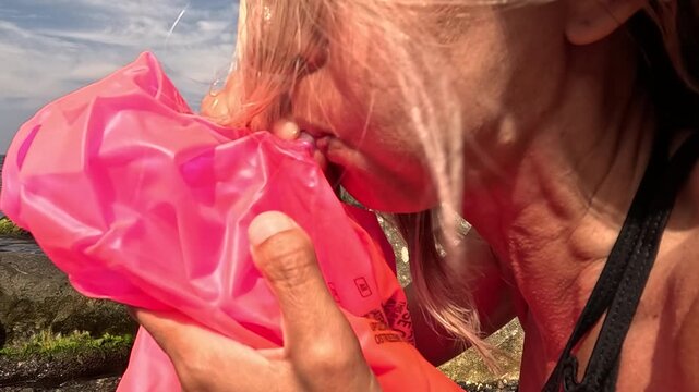 Woman inflates beach toy, Blonde woman blows up pink inflatable on sunny rocky seashore.