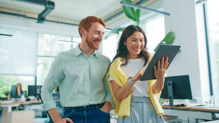Two friendly colleagues standing close together smiling and pointing at tablet screen. Laughing while browsing social media posts. People working in background. Bright workspace with natural daylight. - Powered by Adobe