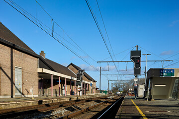 Bright, sunny day at a quiet train station with multiple tracks and platforms under a clear blue sky. Brick station buildings line the left, while modern elements and signal lights mark the right.