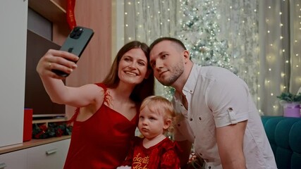 family takes selfie beside christmas tree; white parents and toddler smile into front camera, fairy lights, festive outfits, laughter and warm holiday memories captured - Powered by Adobe