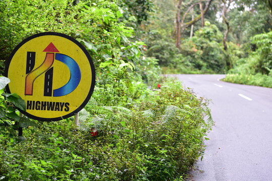 A highway curve warning sign on a scenic forest road in Valparai, Tamil Nadu, surrounded by lush green vegetation during the monsoon season