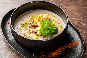 A rich, creamy mushroom soup garnished with croutons, parsley, and spices, served in a black bowl on a rustic wooden table