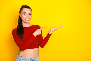 Young woman in red cropped top smiles and points to space against bright yellow background while showcasing casual fashion style