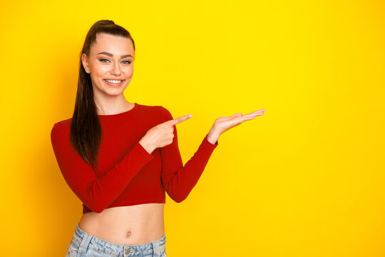 Fototapeta Young woman in red cropped top smiles and points to space against bright yellow background while showcasing casual fashion style