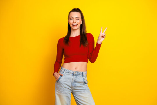 Fototapeta Young brunette woman in a red crop top and jeans smiles against a bright yellow background while posing with a peace sign for a casual lifestyle fashion photo shoot