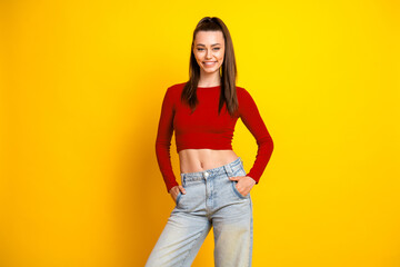 Young brunette woman in red crop top and jeans stands against a yellow background smiling confidently for lifestyle fashion promotion