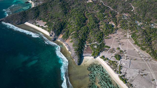 Stunning drone view of a rugged, green forested coastline and cliffs meeting the ocean. Features waves, a white sand beach, and clear, shallow water over a coral reef.