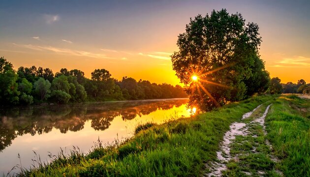 Tranquil river view at sunset with a large tree on the bank. A path winds along the grass
