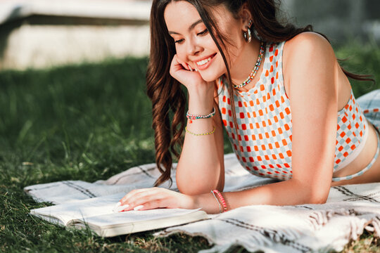 Young woman in a sunny park enjoys a leisure moment reading a notebook on a blanket smiling and relaxing during a casual outdoor day