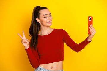 Young woman with red crop top posing for a selfie against a bright yellow background smiling and making a peace sign while holding a red smartphone