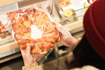 A person in a red hat holds a large, packaged, sweet baked bun or pastry ring, selecting it from a brightly lit supermarket bakery