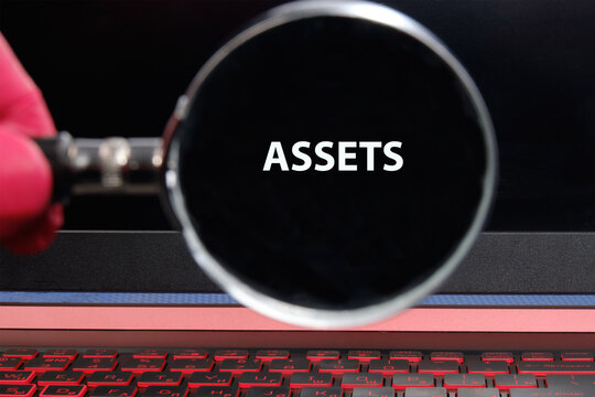 A hand holding a magnifying glass emphasizes the word assets on a laptop screen, symbolizing the importance of identifying valuable resources in the digital age - Powered by Adobe