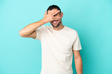 Young handsome caucasian man isolated on blue background covering eyes by hands and smiling
