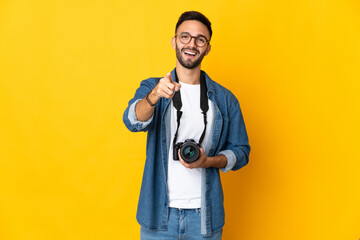 Young photographer girl isolated on yellow background surprised and pointing front