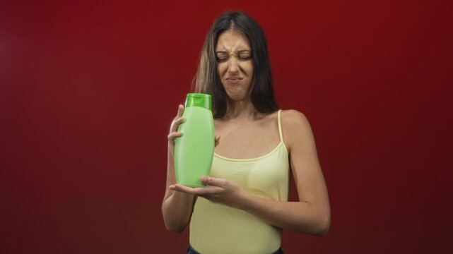 Young woman grimacing while holding green shampoo bottle in red studio; disgust product disappointment.