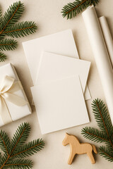 Horizontal composition with white blank cards on a light neutral table. Christmas decor with fir branches, gift box and wrapping paper in soft natural light, Scandinavian minimal style.