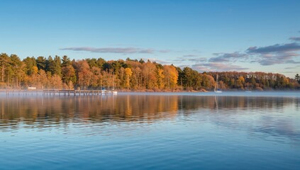 Serene autumn lake at dawn with misty fog rising from water and sailboats docked at a wooden pier surrounded by vibrant fall foliage
