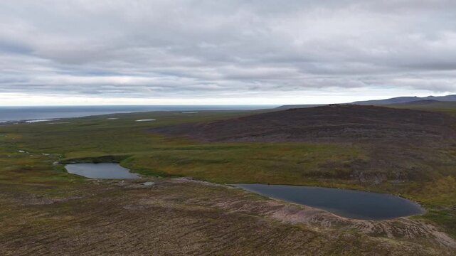 Aerial view of a coastal ridge by Lavrentiya where tundra meets the Bering Sea. Lagoon ponds gleam under a flat cloud ceiling. Polar summer daylight highlights space, wilderness and travel potential