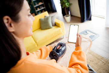 Woman in orange sweater irons at home while using a smartphone in the living room