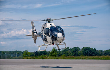 A white Airbus H120 helicopter hovers just above the runway, landing skids extended, rotor in motion, framed against trees and a layered sky filled with alto-stratus and cirrus cloud formations. © Paul