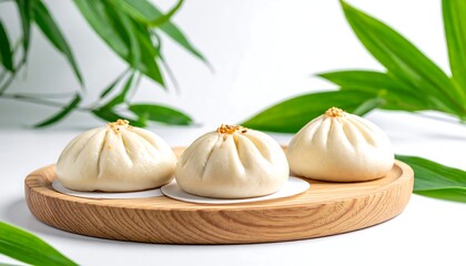 Three steamed buns sit on a round wooden plate with green leaves against a white backdrop creating a minimalist scene