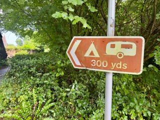 A brown tourist information road sign indicates the direction of a campsite .Arrow points left. White symbols on brown background.Set in countryside.