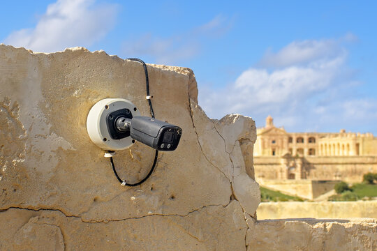 Modern surveillance camera mounted on a weathered stone wall with a historic building in the background under blue sky.
