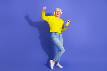 Energetic cheerful mature female dancing with headphones holding phone against purple background for lifestyle advertising