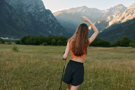 A young woman stands in a wide alpine meadow, facing distant mountains as she raises her arm to shade her eyes, enjoying a sunny hike and fresh air.