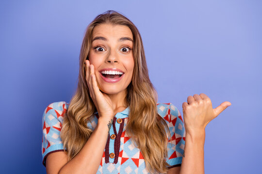 Joyful young woman with bright smile gestures and pointing against a blue background wearing a colorful geometric sweater - Powered by Adobe