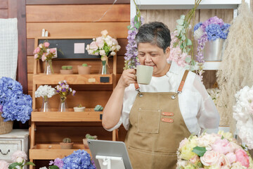 Asian senior florist sipping warm tea with closed eyes enjoying peaceful moment during break in cozy flower shop filled with pastel bouquets and floral decor after morning customer orders